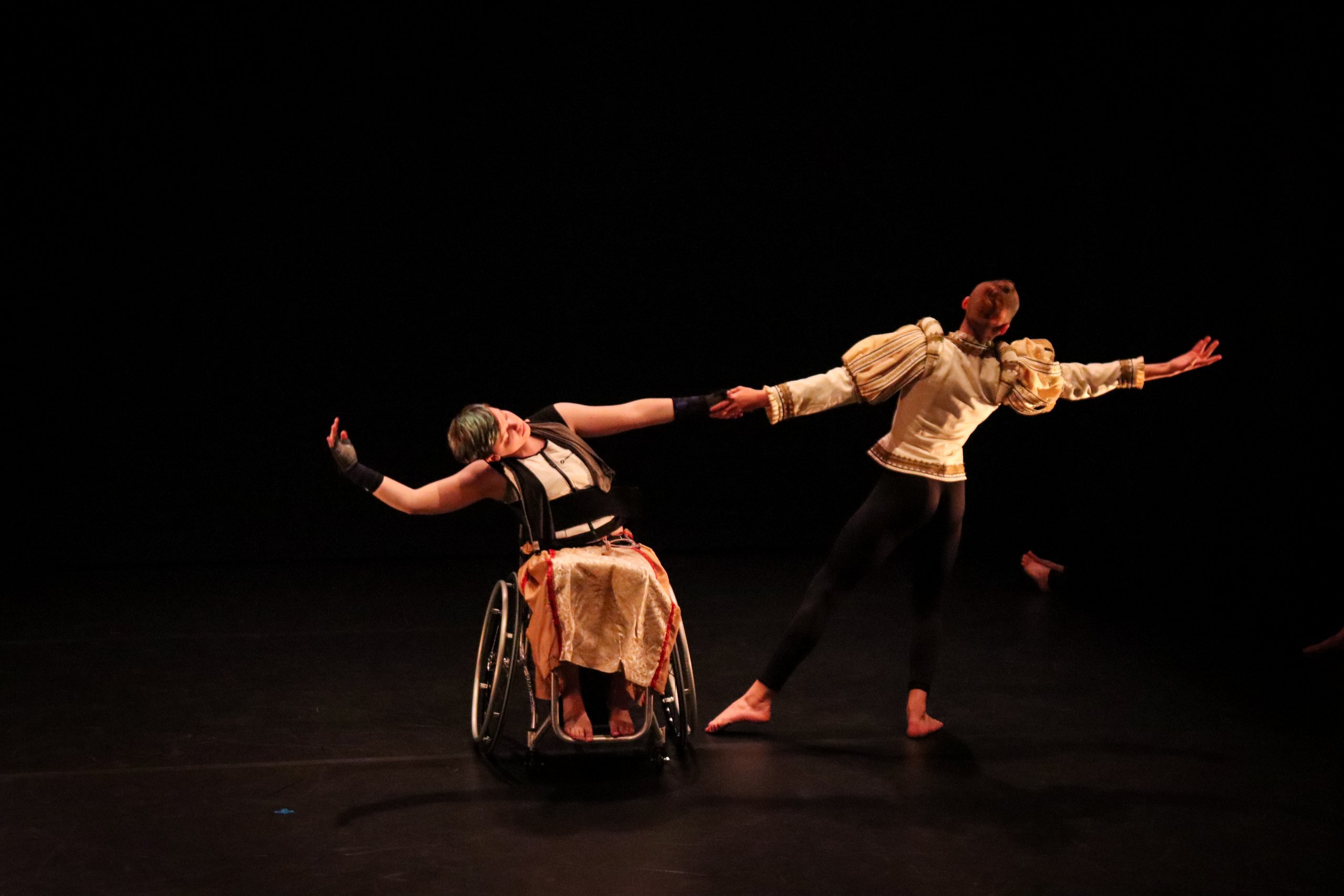 A white woman in a wheelchair dancing with a male performer. The background is black